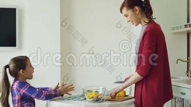 可爱的女儿和开朗的母亲在现代厨房做饭时交谈。 妈妈用一块蔬菜喂她女儿